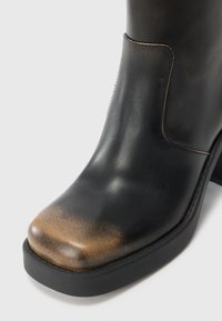 Bottillon en cuir noir avec un bout arrondi, présentant un accent en bronze dégradé sur le capuchon, et une semelle en caoutchouc noir robuste.
