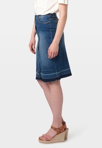 Falda de mezclilla en forma de A en azul oscuro con cremallera vertical, costuras en contraste y dobladillo a la altura de la rodilla. Llevada con sandalias de cuña con tiras marrones.