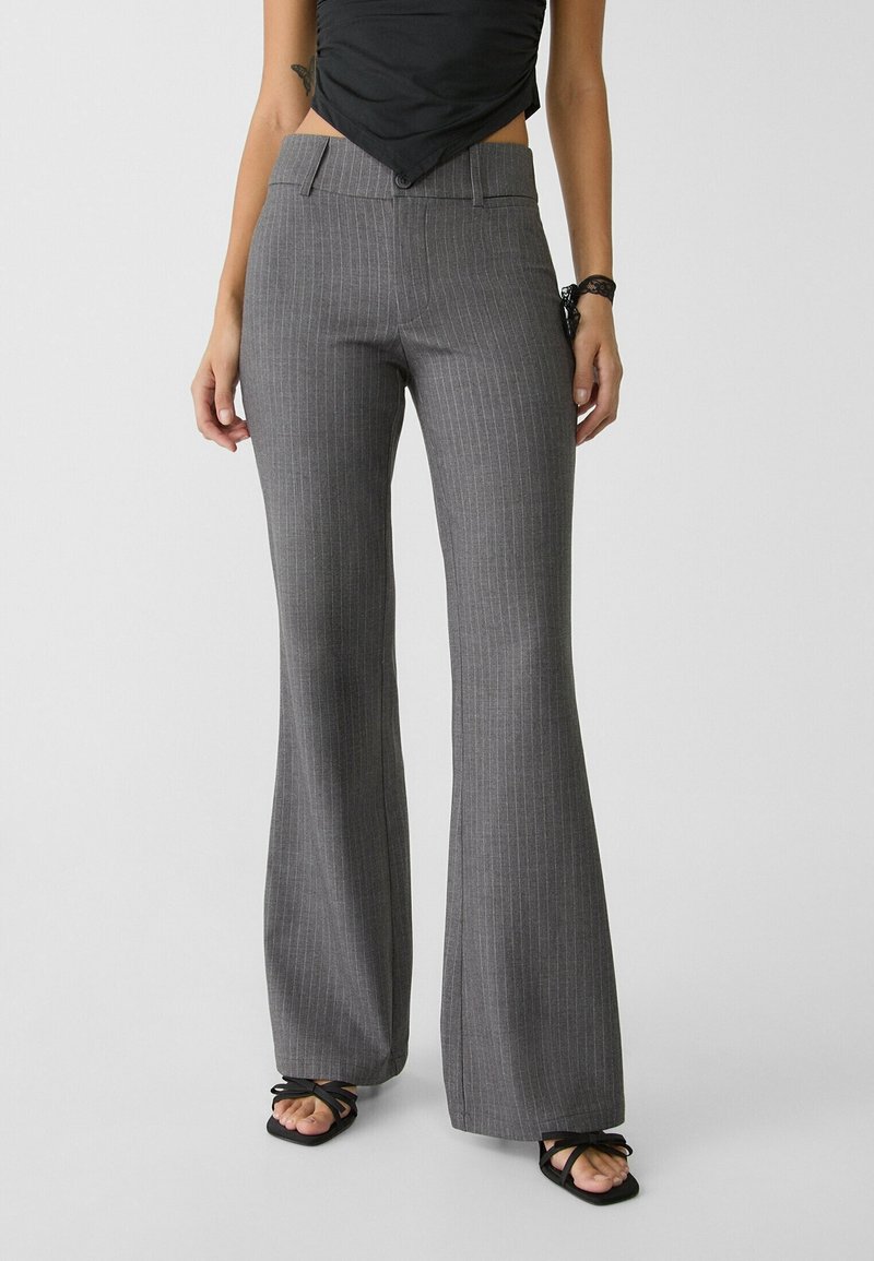 Femme portant un pantalon gris à fines rayures taille haute évasé et des sandales noires à lanières et bouts ouverts, debout devant un fond uni.