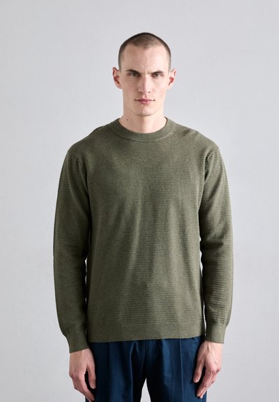 Pull en tricot vert olive avec un col rond et des manches longues, présentant un tissu texturé et des poignets et un ourlet côtelés. Associé à un pantalon bleu.