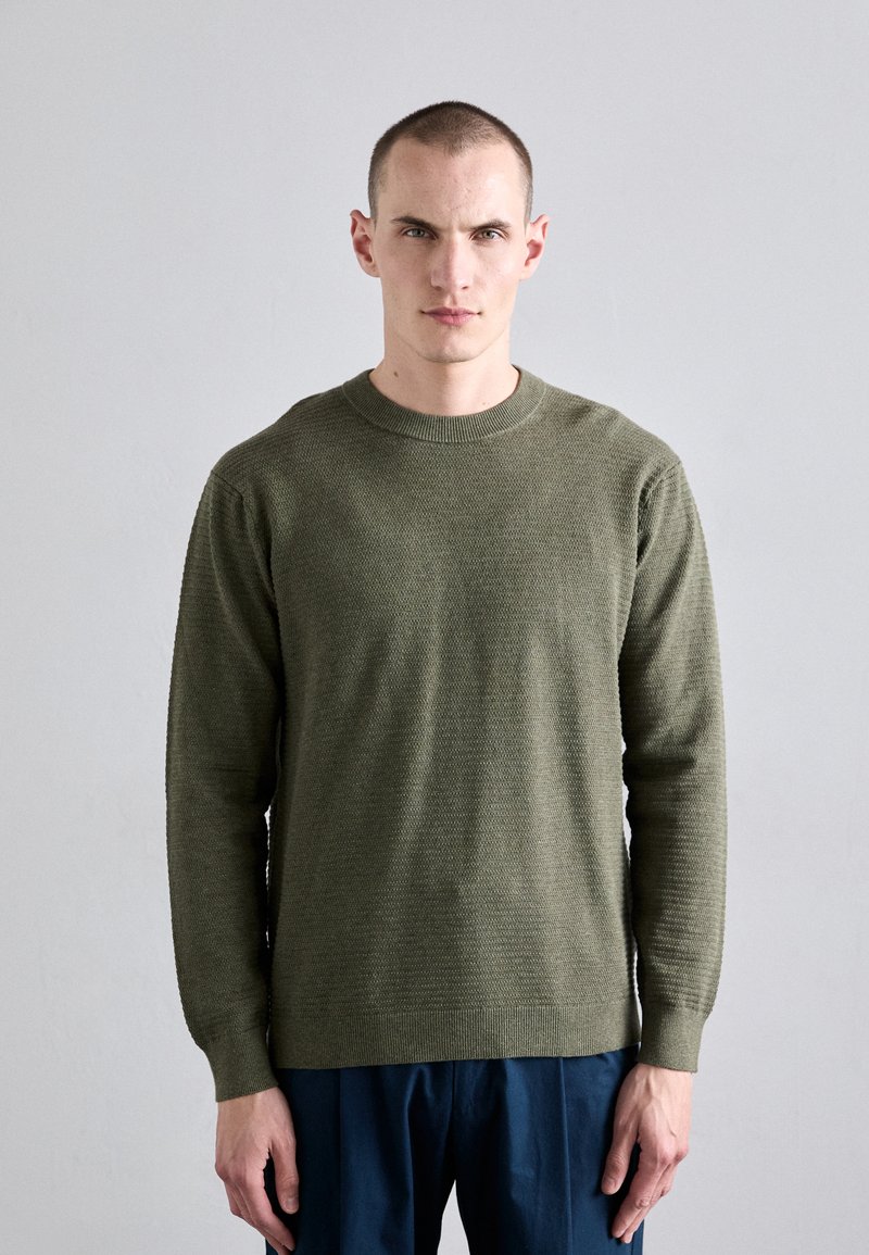 Pull en tricot vert olive avec un col rond et des manches longues, présentant un tissu texturé et des poignets et un ourlet côtelés. Associé à un pantalon bleu.
