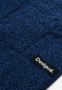 Blue textured fabric with a woven pattern and a small black tag labeled "Desigual." featuring a rectangular pocket detail.