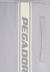 Ljust grå klädsel med ett vertikalt vitt rand med "PEGADOR"-text i grått. Tyget är slätt med en sidofickdetalj.