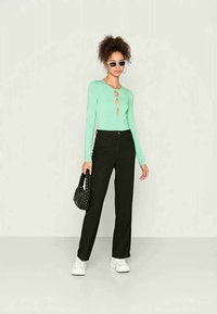 Blusa de manga longa verde-menta com detalhe em forma de chave, combinada com calças pretas de cintura alta e ténis brancos. Bolsa trançada preta e óculos de sol.