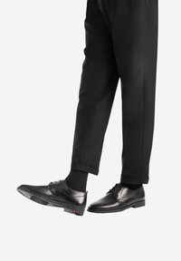 Scarpe eleganti in pelle nera con una finitura liscia, lacci e dettagli testurizzati, abbinate a pantaloni neri, che mostrano una vestibilità su misura.
