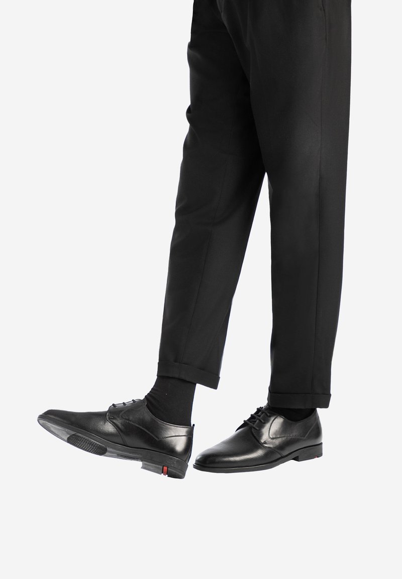 Scarpe eleganti in pelle nera con una finitura liscia, lacci e dettagli testurizzati, abbinate a pantaloni neri, che mostrano una vestibilità su misura.