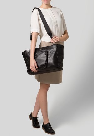Femme portant un chemisier blanc et une jupe beige, portant un grand sac fourre-tout en cuir noir avec une bandoulière et des poignées.