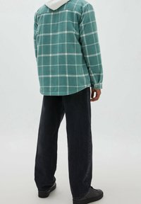 Persona in piedi con le spalle alla telecamera, che indossa una camicia a quadri turchese sopra un felpa bianca e pantaloni neri, su uno sfondo neutro.