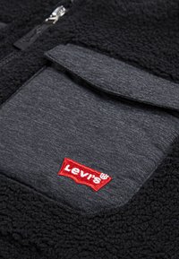 Fleecejakke med en svart teksturert kropp og glatt grå panel, med en rød brodert "Levi's"-logo på framlommen.