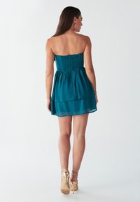Teal strapless jurk met een smock-borstvoordeel, gelaagde rok en een doorschijnende overlay. Bevat strappy gouden hakken met een bloemens accent.