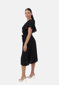 Vestido negro con mangas cortas y ligeras, y cintura ceñida con cinturón. Textura suave, a la altura de la rodilla, combinado con mule de tacón de color claro.