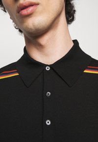 Czarna koszulka polo z klasycznym kołnierzykiem, trzema czarnymi guzikami oraz czerwonymi, żółtymi i niebieskimi paskami na ramionach.