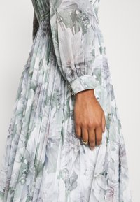Femme portant une robe longue légère à manches longues à motif floral avec des plis, montrant un gros plan de son bras et de sa main avec deux bagues en or.