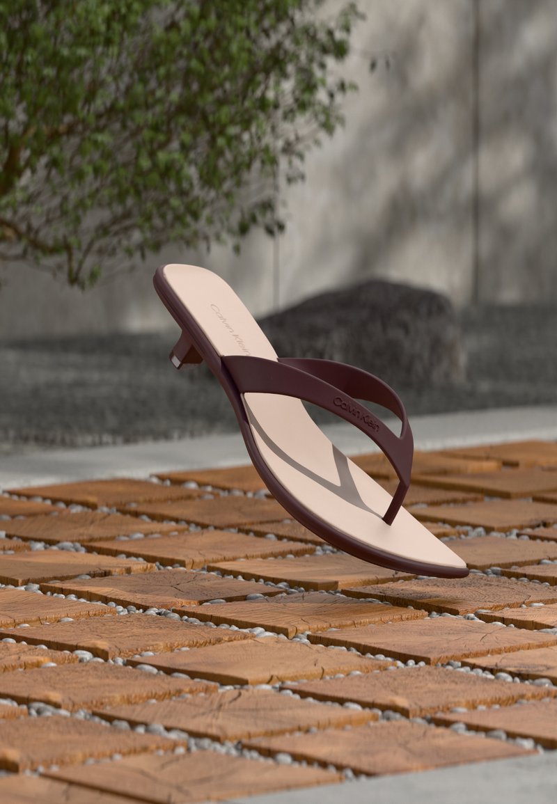 Μπεζ σανδάλι με σκουρό καφέ λουριά και χαμηλό τακούνι που αιωρείται πάνω από μια ξύλινη επιφάνεια με σχέδια, σε εξωτερικό χώρο με πράσινο και πέτρινο τοίχο στο φόντο.