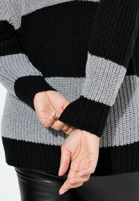 Gestrickter Pullover mit schwarzen und grauen horizontalen Streifen. Verfügt über gerippte Bündchen und eine strukturierte Optik. Die Hände des Modells sind hinter dem Rücken verschränkt.