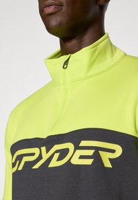 Gul og grå zip-up genser med høy krage, myk tekstur og stor "SPYDER"-logo på brystet i fet gul skrift.