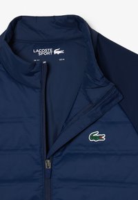 Giacca trapuntata blu navy con collo alto, cerniera frontale e logo del coccodrillo Lacoste. Presenta una texture liscia e un materiale leggero.