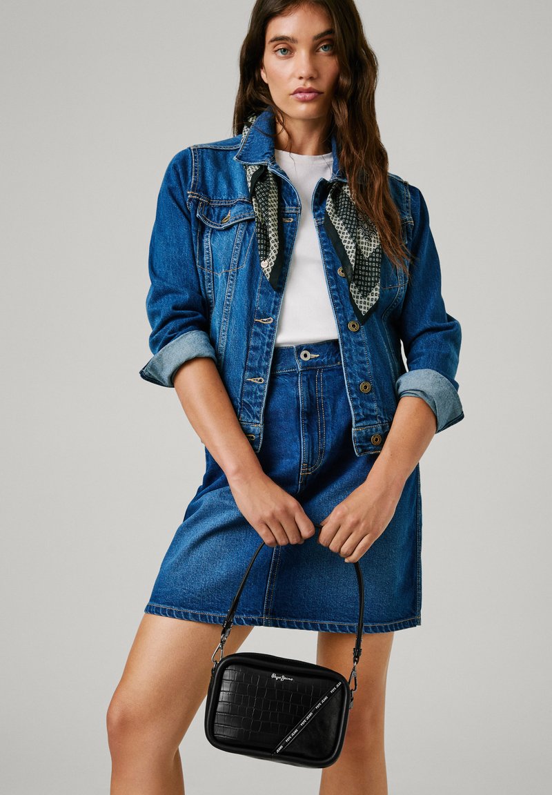 Blauwe jeansjacket, witte top en blauwe rok. Zwarte, gestructureerde handtas met een dunne schouderband die voorgehouden wordt. Sjaal om de kraag van de jacket.