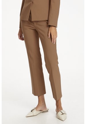Femme portant un pantalon marron court et ajusté ainsi que des chaussures blanches sans lacets à bouts pointus, debout sur un fond blanc.