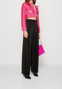 Mujer con una chaqueta corta de felpa rosa, pantalones de pierna ancha negros de cintura alta, tacones negros de punta y sosteniendo un bolso de peluche rosa.
