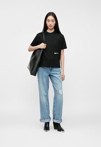 Ung kvinne iført svart t-skjorte, lyse ripped jeans, svarte ankelstøvler, bærende en stor svart tote veske på skulderen, stående.