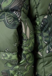 Chaqueta de abrigo verde con estampados de dinosaurios de dibujos animados, material de nylon brillante, diseño acolchado y cierre de botones a presión. Textura suave.