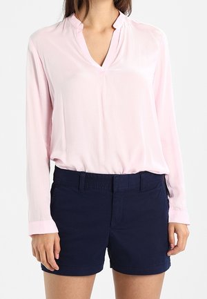 Femme portant un chemisier à manches longues rose clair rentré dans un short bleu marine ajusté, sur un fond uni.