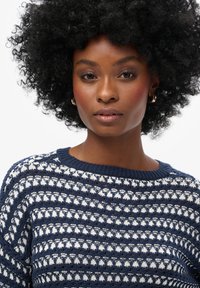 Joven mujer con cabello rizado natural, que lleva pendientes de aro dorados y un suéter de punto azul y blanco con un patrón, mirando hacia adelante con confianza.