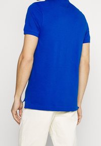 Sininen puuvillapolo paita lyhyillä hihoilla, litteällä kauluksella ja sivuhaaroilla helman kohdalla, yhdistettynä vaaleisiin housuihin.