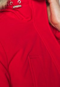 Chaqueta roja con capucha de textura suave, que cuenta con cordón de ajuste y un bolsillo frontal tipo parche. El diseño enfatiza una silueta moderna y elegante.