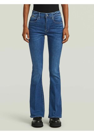 Persona che indossa jeans svasati a vita alta blu, scarpe nere e top nero, in piedi davanti a uno sfondo neutro.