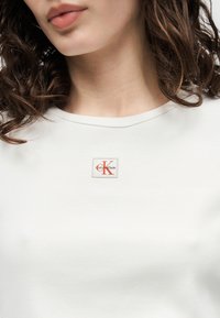 Γυναίκα με καστανά σγουρά μαλλιά που φοράει λευκό ριμπ τοπ Calvin Klein με κόκκινο λογότυπο "CK" κεντραρισμένο κάτω από το λαιμό.