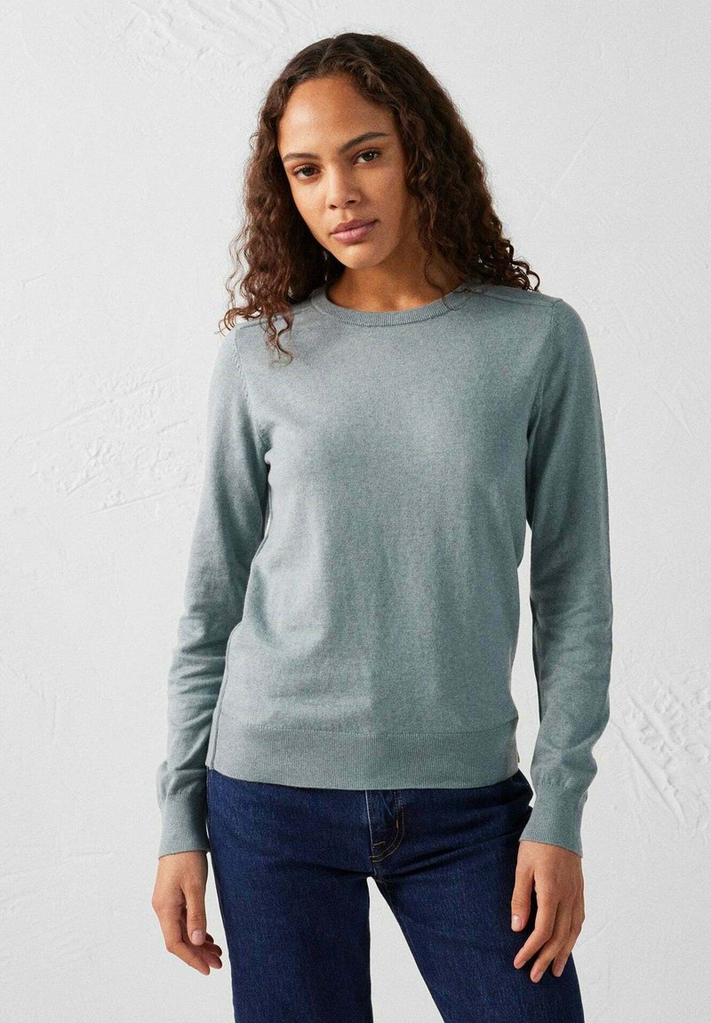 Lichtgrijze trui van zachte stof, met een ronde hals, lange mouwen en ribgebreide manchetten. Gepaard met donkerblauwe jeans.
