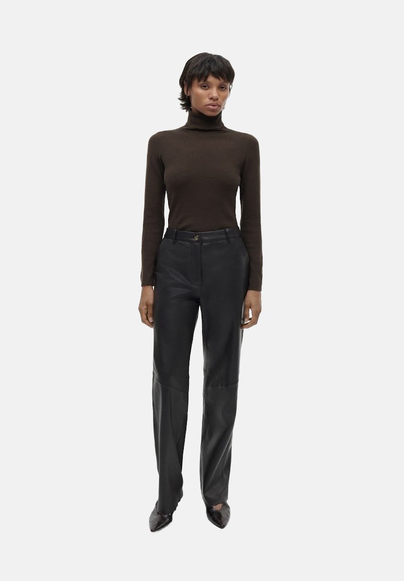 Brauner Rollkragenpullover und schwarze Lederhose. Die Hose hat einen geraden Schnitt mit Einsätzen. Das Modell trägt spitze schwarze Schuhe.
