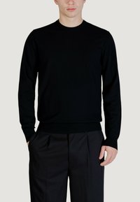 Maglione nero girocollo in tessuto morbido con maniche lunghe e polsini a coste. Abbinato a pantaloni neri su misura con pieghe.