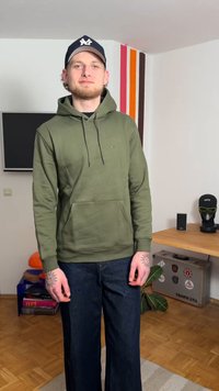 Sweat à capuche vert olive avec poche avant et cordons. Jean noir, coupe décontractée. Tatouages visibles sur les bras, portant une casquette sombre.