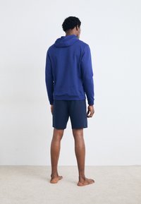 Marineblaue Kapuzenjacke mit Kordelzug und gerippten Bündchen, kombiniert mit passenden marineblauen Shorts, von hinten gesehen. Weiche Stofftextur.