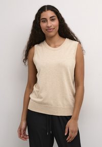 Haut tricot beige sans manches avec un col rond et un ourlet côtelé. Le tissu semble doux avec une texture subtile. Porté avec un pantalon noir.