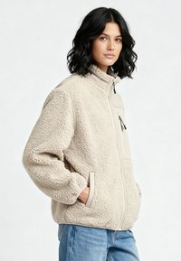 Veste polaire beige avec col montant, poches zippées et accents contrastants aux épaules. Présente une surface douce et texturée ainsi qu'une coupe décontractée.