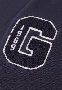 Námořnicky modrá látka s texturovaným bílým a tmavě modrým vyšívaným detailem „G“ a „1969“. Hladká textura, detailní záběr designových prvků.