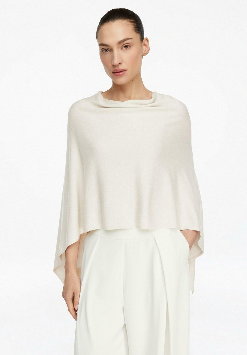 Woman with dark hair tied back wearing a loose, cream-colored poncho and high-waisted white trousers, standing with one hand in pocket.