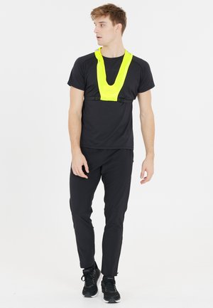 Sort atletisk skjorte, sorte bukser og sorte sneakers. Indeholder en klar gul refleksvest med et V-formet design og justerbare stropper.