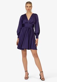 Abito di raso viola con scollo a V profondo, maniche lunghe a sbuffo e vita arricciata. La gonna si allarga, presentando una texture liscia e un'elegante caduta.