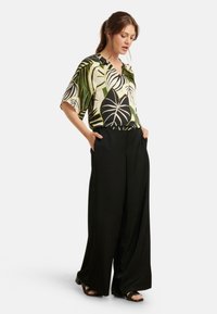 Kurzärmlige Bluse mit tropischem Blattdruck in Schwarz, Grün und Creme. Breite schwarze Hose, glattes Material und lockere Passform.