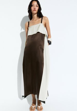 Donna con lunghi capelli scuri che indossa un abito maxi senza maniche marrone e crema a blocchi di colore e sandali bianchi, in piedi davanti a uno sfondo bianco uniforme.