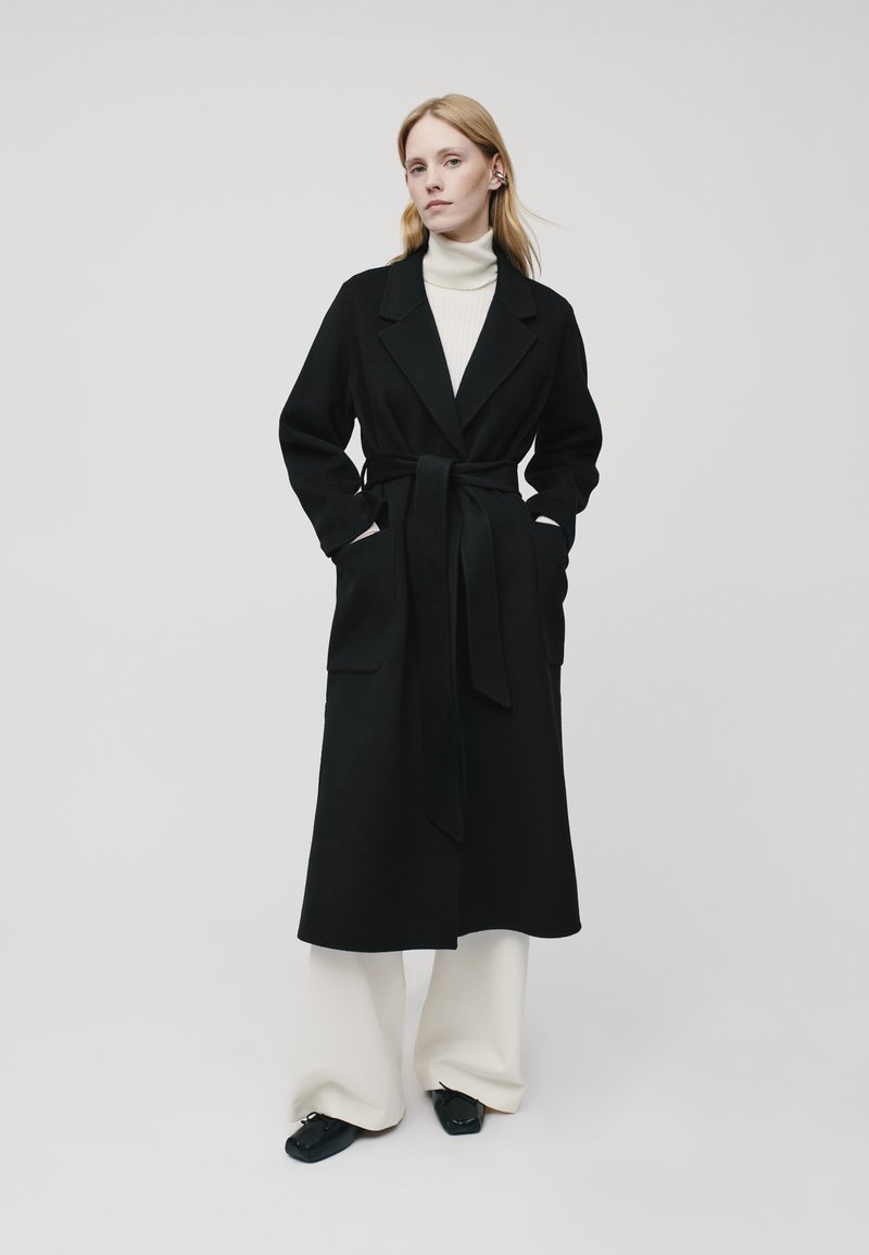 Black long coat with a tie belt, wide lapels, and side pockets, paired with a light turtleneck and wide cream pants, smooth texture.