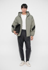 Chaqueta tipo puff en verde oliva con capucha y detalles en negro, combinada con pantalones deportivos negros con rayas laterales blancas. Zapatillas beige.