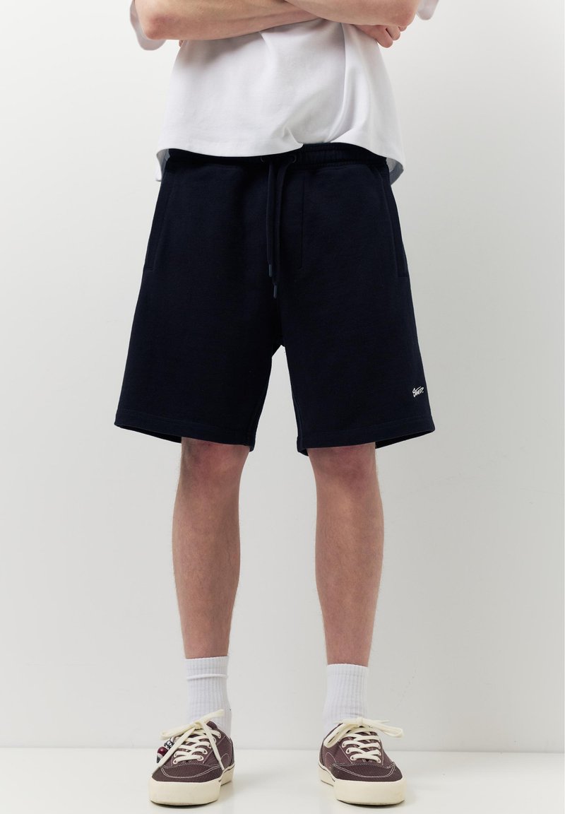 Personne portant un short noir à cordon, des chaussettes blanches et des baskets marron, debout les bras croisés devant un fond blanc uni.