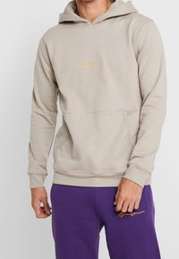 Beige mikina s kapucí z bavlněné směsi s přední kapsou. Na hrudi se nachází zlatá výšivka loga. Dlouhé rukávy, normální střih.