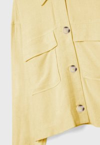 Chemise en lin jaune à coupe décontractée, dotée de deux grandes poches poitrine et de boutons arrondis pour la fermeture. Tissu texturé avec un aspect naturel.